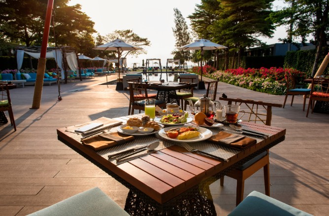 http://A%20sunlit%20outdoor%20dining%20setting%20with%20a%20wooden%20table%20featuring%20a%20variety%20of%20breakfast%20dishes.%20The%20background%20shows%20a%20pool,%20umbrellas,%20and%20lush%20greenery.