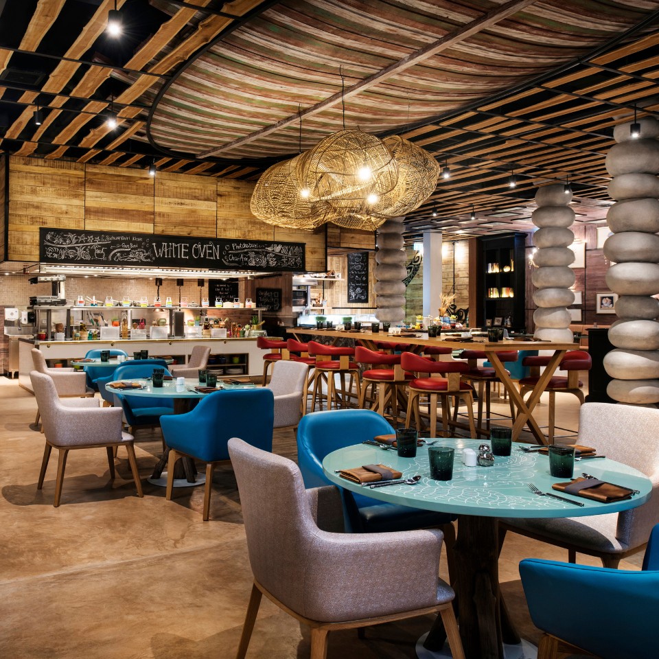 Modern restaurant interior with a warm, inviting ambiance. Features stacked stone columns, turquoise tables, beige and red chairs, and woven light fixtures.