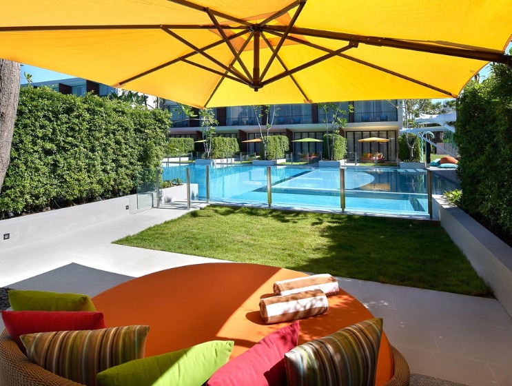 Outdoor scene with a large yellow umbrella shading a round table with colorful cushions. A pool enclosed by glass fencing lies beyond a grassy area, surrounded by lush greenery and modern buildings, creating a tranquil, inviting atmosphere.