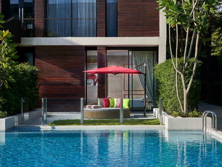 Modern three-story building with wooden facade, large windows, and a serene pool. A red umbrella and greenery add vibrancy and a relaxing tone.