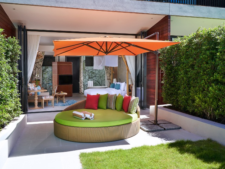 Spacious patio with a circular daybed, vibrant green and red cushions beneath an orange umbrella. Beyond, a cozy room opens with gentle, earthy tones.