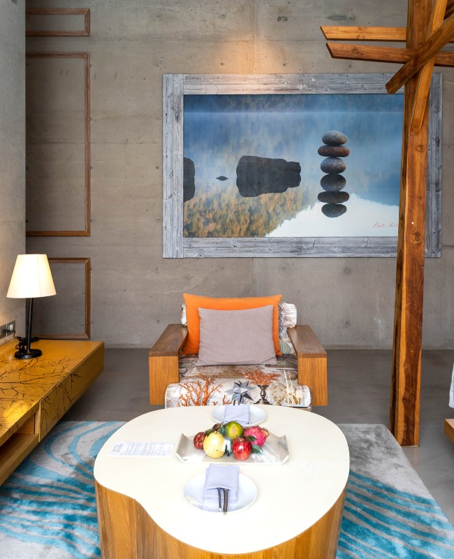 A cozy modern room with a wooden chair, vibrant cushions, and a table topped with fruit. A serene painting of stacked stones and water hangs on a concrete wall.