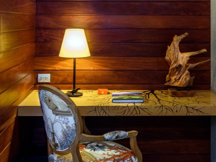 Cozy study nook with warm lighting. A wooden desk features a tree design, adorned with a driftwood sculpture and books. A patterned chair adds elegance.