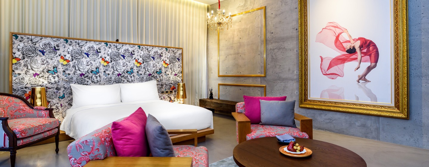 Luxurious hotel room with a large bed, floral and butterfly headboard, pink and gray ornate chairs, gold accents, and a chandelier, creating an elegant atmosphere.