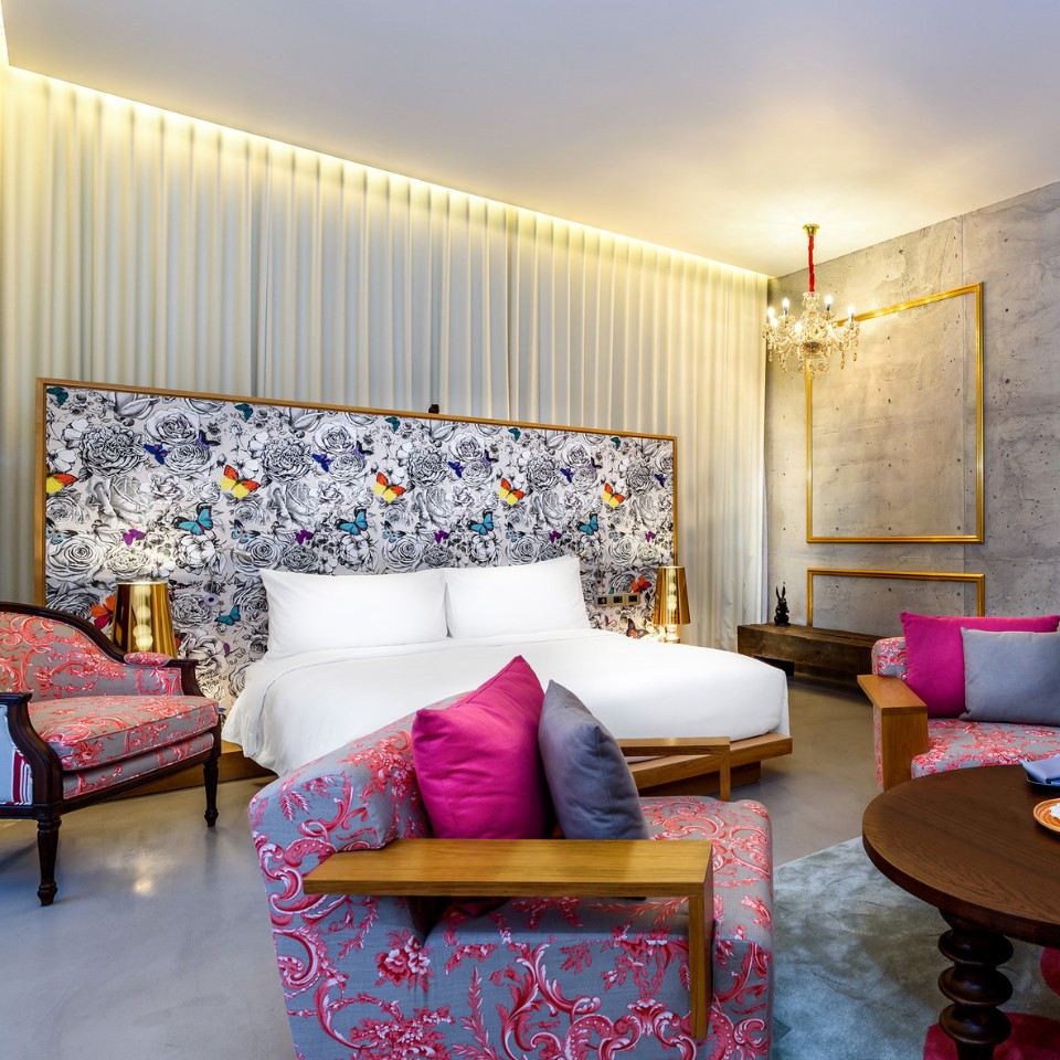Luxurious hotel room with a large bed, floral and butterfly headboard, pink and gray ornate chairs, gold accents, and a chandelier, creating an elegant atmosphere.