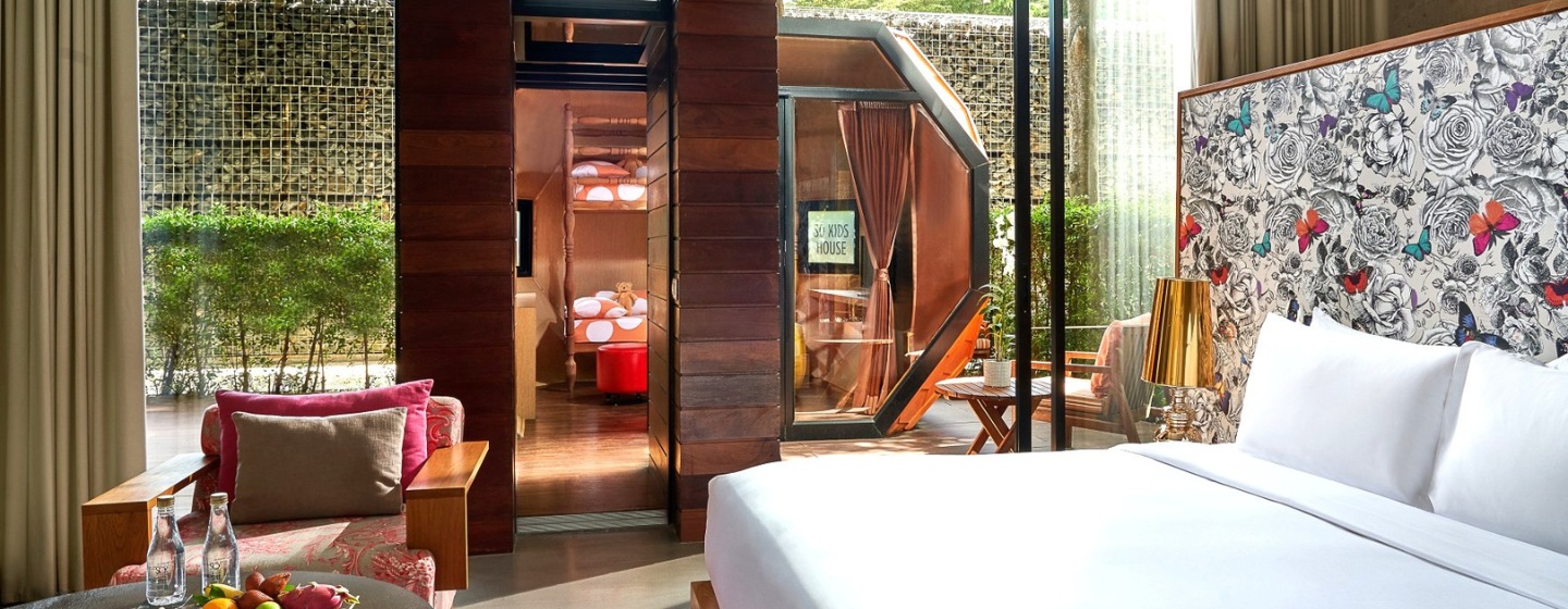Modern hotel room with a large bed, patterned butterfly wallpaper, and a cozy seating area. Glass doors lead to a lush outdoor patio, creating a serene ambiance.