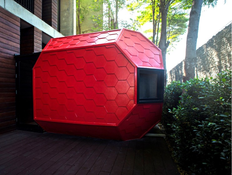 Red hexagonal pod with a window, attached to a modern building with wooden walls. Surrounded by greenery and trees, it creates a futuristic, cozy vibe.
