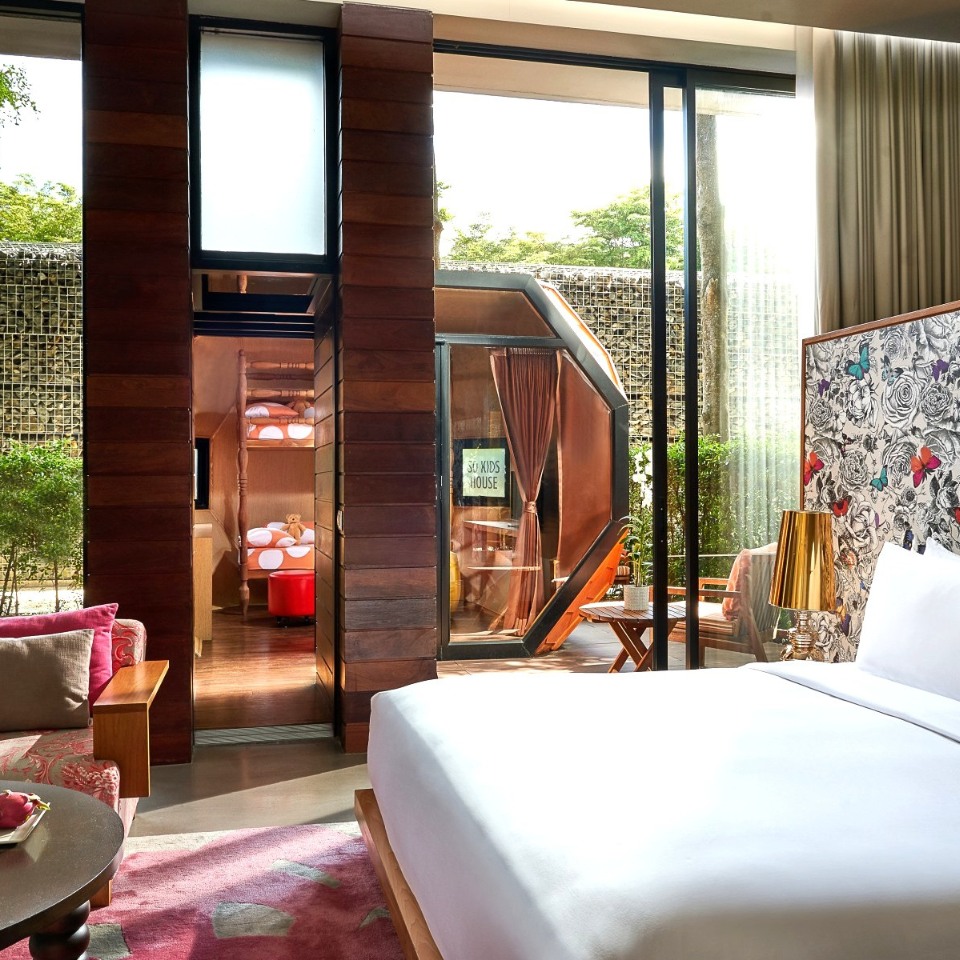 Modern hotel room with a large bed, patterned butterfly wallpaper, and a cozy seating area. Glass doors lead to a lush outdoor patio, creating a serene ambiance.
