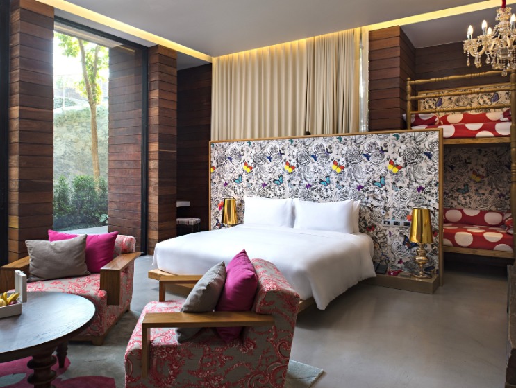 Modern hotel room with a vibrant floral headboard, white linens, wooden accents, and colorful cushions. A chandelier and bunk beds add elegance and playfulness.