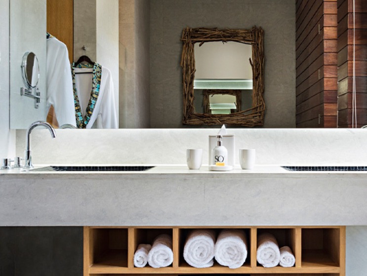Modern bathroom with a large white countertop and dual sinks. Above, a rustic mirror with a driftwood frame. Rolled towels in cubbies, robe hanging nearby.