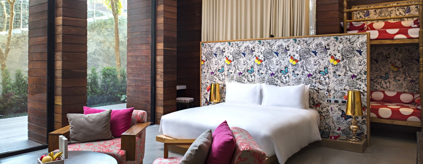 Modern hotel room with a large white bed, decorative floral wall, and playful bunk beds. Warm, wooden accent walls and bright pink cushions create a cozy, welcoming ambiance.