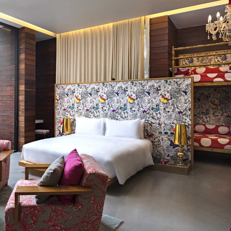 Modern hotel room with a large white bed, decorative floral wall, and playful bunk beds. Warm, wooden accent walls and bright pink cushions create a cozy, welcoming ambiance.