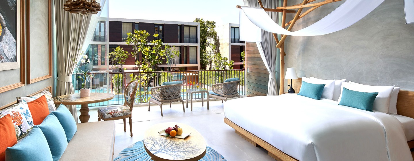 Bright, modern hotel room featuring a king bed, lounge area, and a balcony overlooking greenery and a swimming pool.