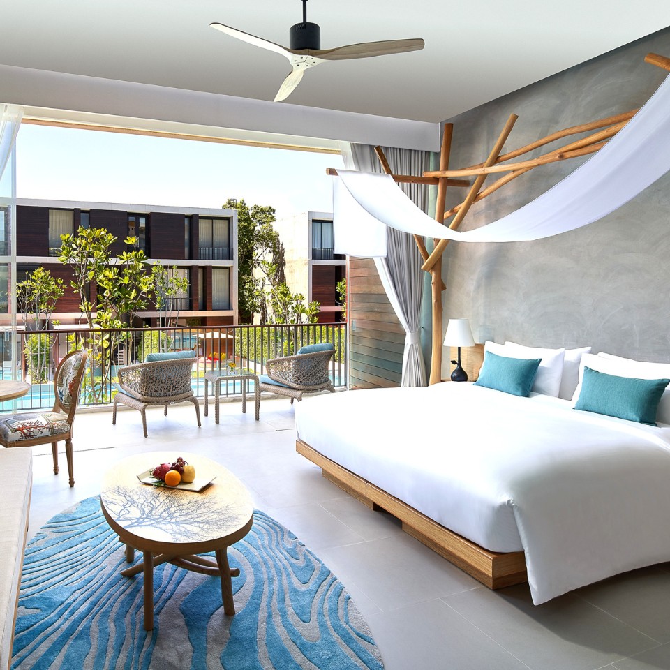Bright, modern hotel room featuring a king bed, lounge area, and a balcony overlooking greenery and a swimming pool.