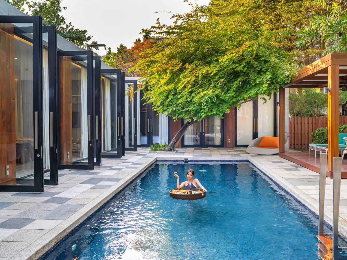A woman relaxes on a donut-shaped float, enjoying food in a modern pool. Glass doors and lush trees create a serene, luxurious ambiance.