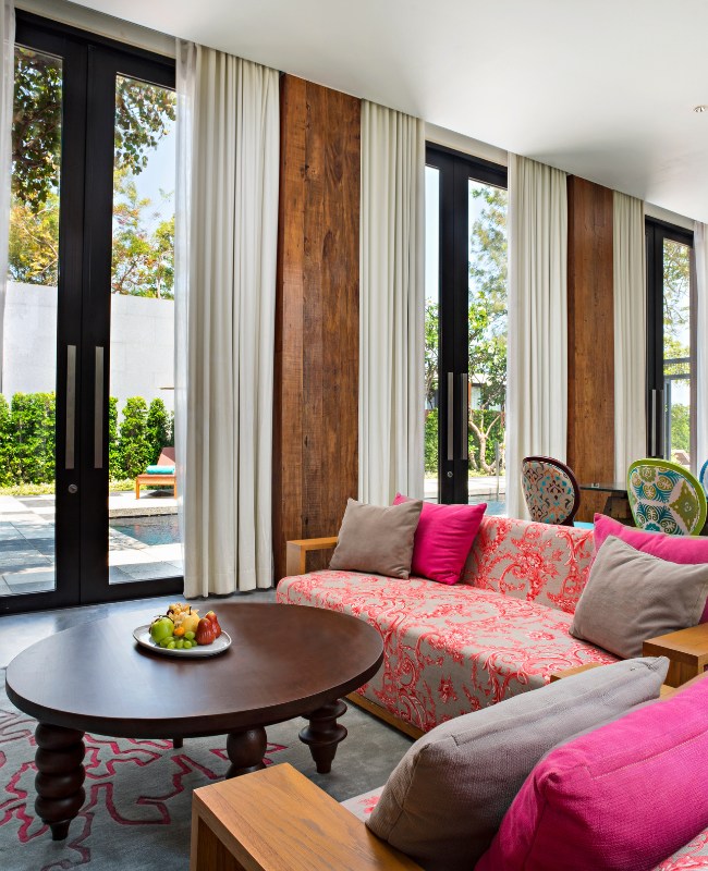 Cozy living room with large windows and white curtains. Features a pink floral sofa with colorful cushions, a wooden coffee table, and a serene outdoor view.