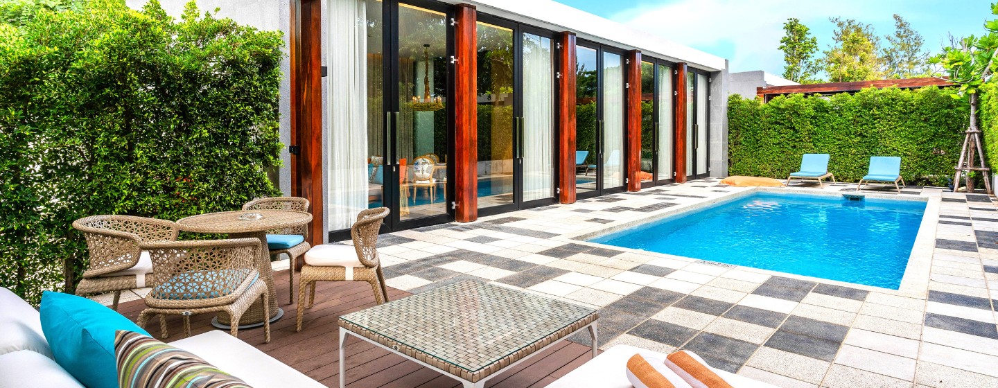 Luxurious poolside patio with a modern square pool surrounded by sun loungers, greenery, and glass doors. Bright, sunny day, relaxing ambiance.