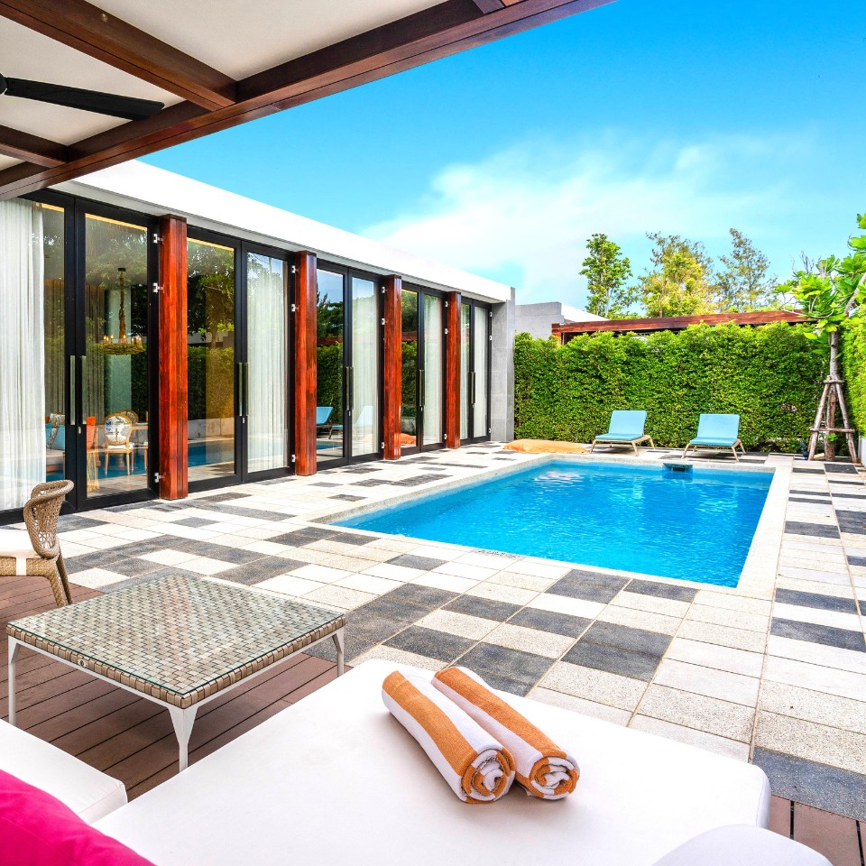 Luxurious poolside patio with a modern square pool surrounded by sun loungers, greenery, and glass doors. Bright, sunny day, relaxing ambiance.