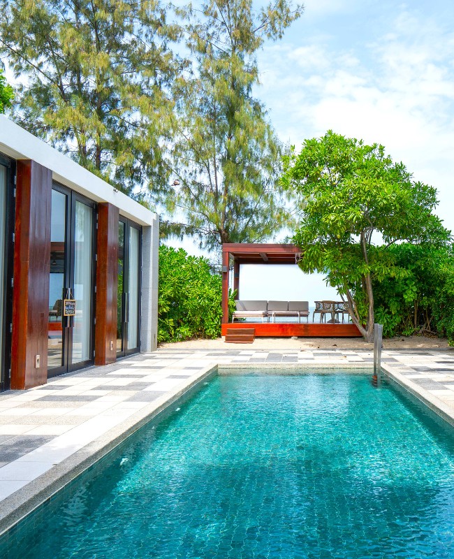 A serene outdoor scene featuring a turquoise pool beside a modern house with glass doors. A cozy patio area and lush greenery enhance the tranquil vibe.