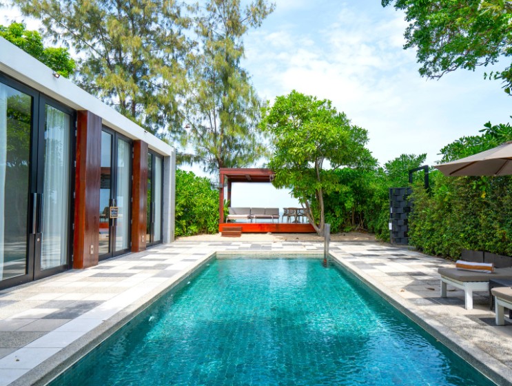 A serene outdoor scene featuring a long, rectangular swimming pool lined with trees and lounge chairs under umbrellas. Large glass doors open onto the area.