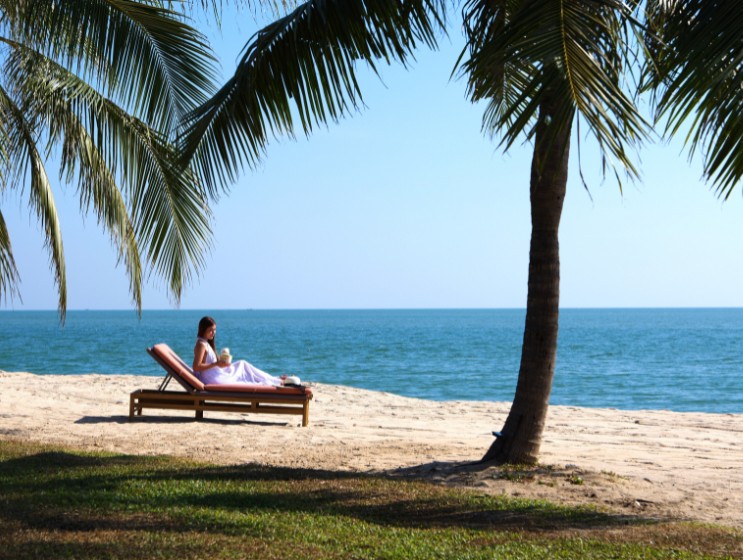 A woman lounges on a beach chair under palm trees, gazing at the serene ocean. The scene conveys relaxation and tranquility on a sunny day.