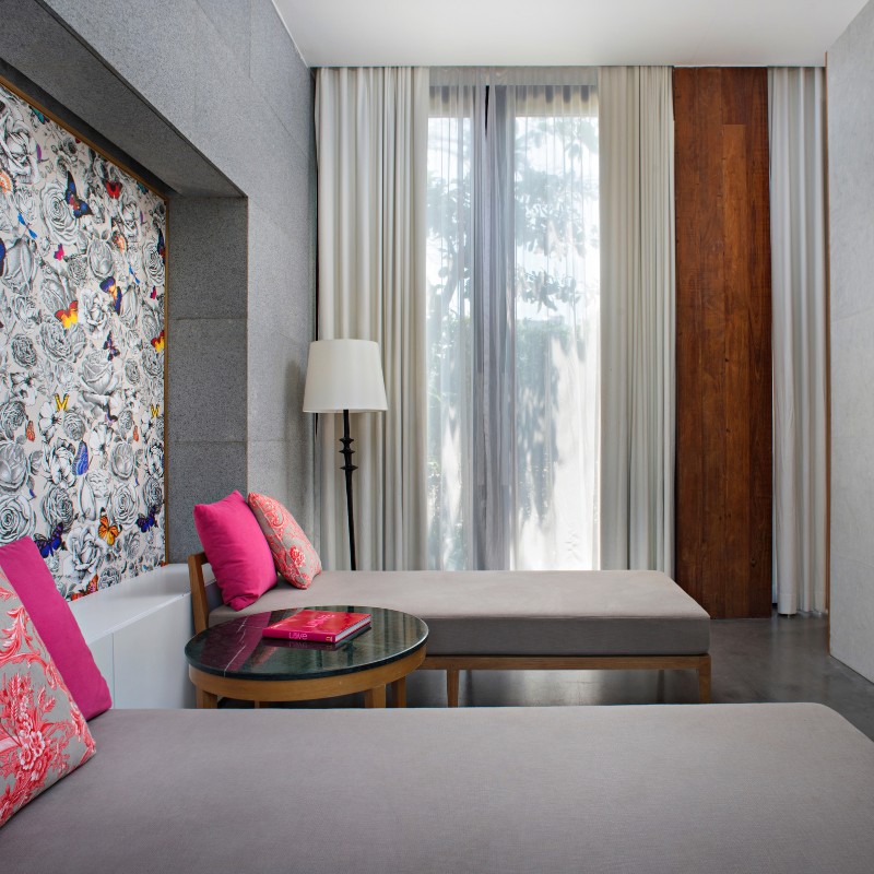 Modern room with two gray daybeds featuring pink cushions. A floral and butterfly mural adorns one wall. A sheer-curtained window and tall lamp add light.