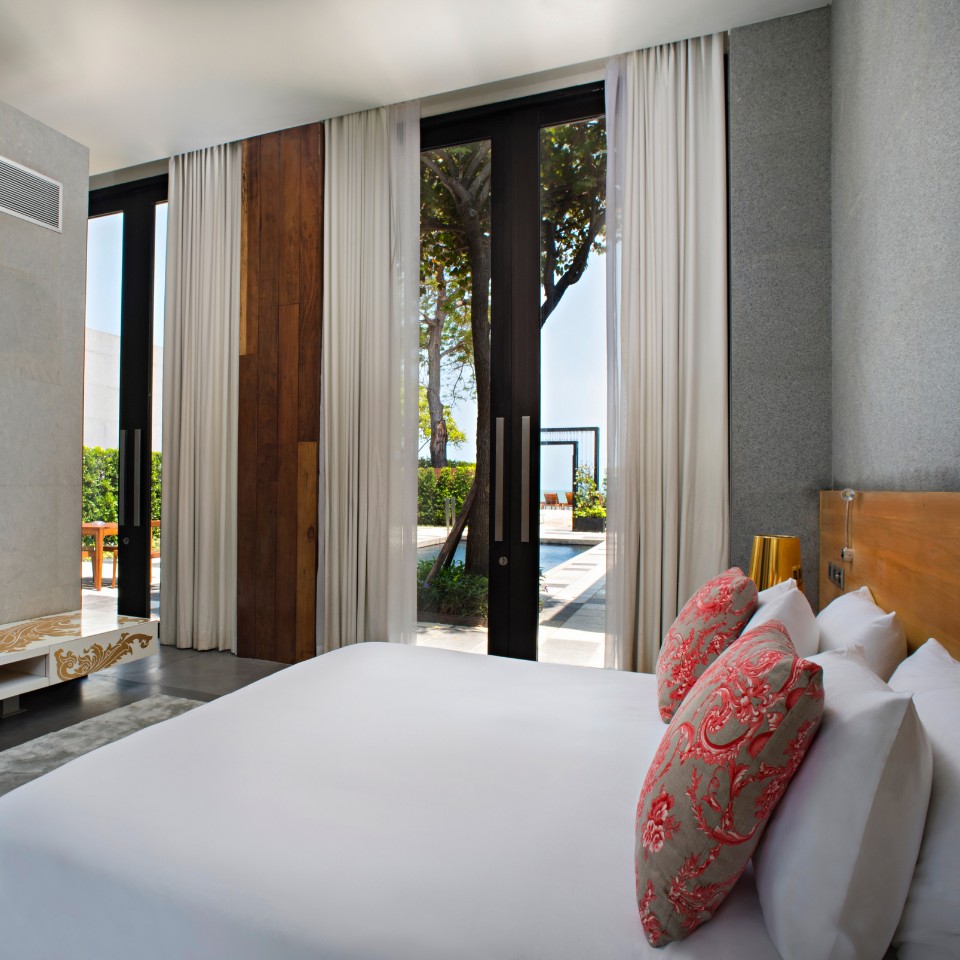 Modern bedroom with a large bed adorned with red-patterned pillows. A wall-mounted TV sits opposite. Floor-to-ceiling windows reveal a sunny patio.