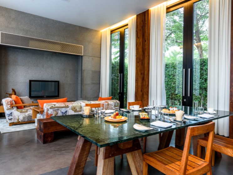 Contemporary dining room with a glass table set for six. Wooden chairs, a modern sofa with orange cushions, and large windows create a bright, inviting atmosphere.
