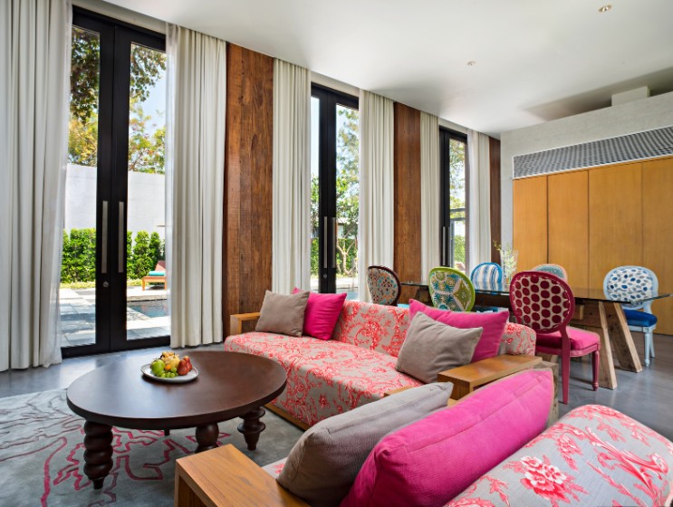 Modern living room with large windows, light curtains, pink floral sofas, round wooden table with fruit plate, and colorful, chic dining chairs.