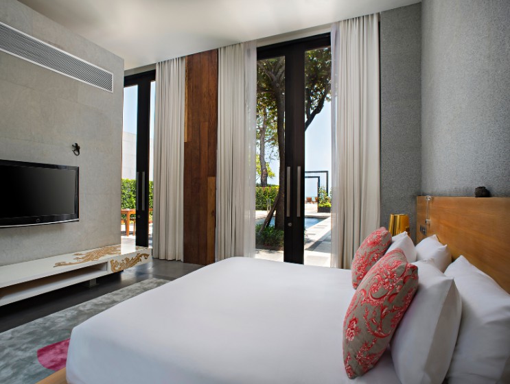 Modern bedroom with large windows, white curtains, and garden view. Features a stylish bed with red-patterned pillows, wall-mounted TV, and wood accents.