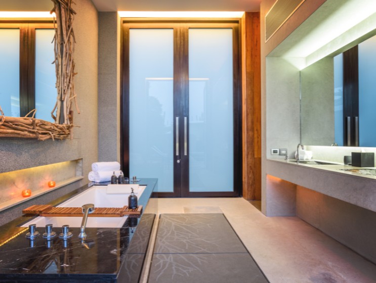 Modern bathroom with a sleek black tub, wooden décor accents, and frosted glass doors. Warm lighting and candles create a serene, spa-like ambiance.