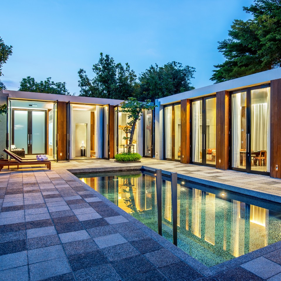 Modern house exterior at dusk with tall glass doors and warm lighting. A sleek pool reflects the building, surrounded by trees, conveying tranquility and luxury.