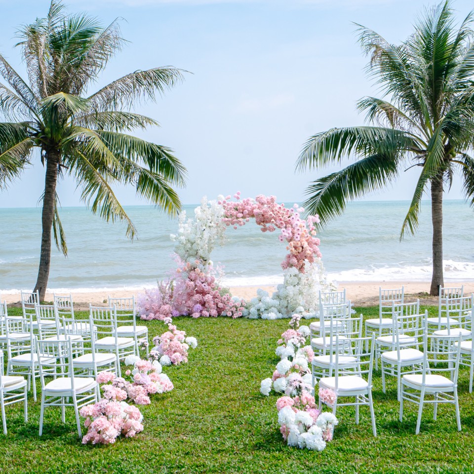 Beach wedding setup with white chairs arranged in rows facing a floral arch adorned with pink and white flowers. Palm trees frame the scene, under a clear blue sky.