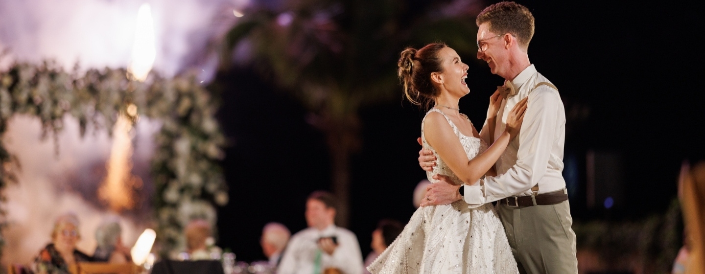 A smiling couple shares a joyful dance at their wedding, surrounded by lush greenery and elegant decorations. Fireworks light up the night sky.