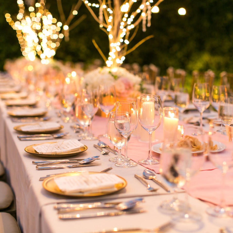 Elegant outdoor dining setup with a long table adorned with pink flowers, candles, and glassware, creating a warm, romantic atmosphere. Soft lighting enhances the mood.