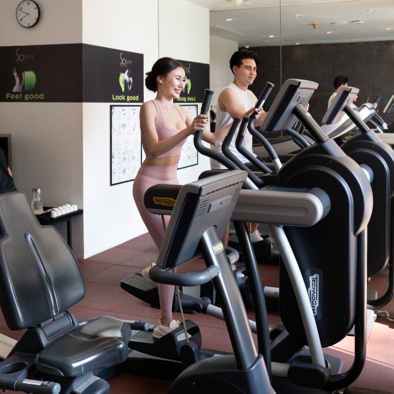 Two people exercise on elliptical machines in a modern gym. They appear focused and fit. A motivational "Feel good, look good" sign is visible.