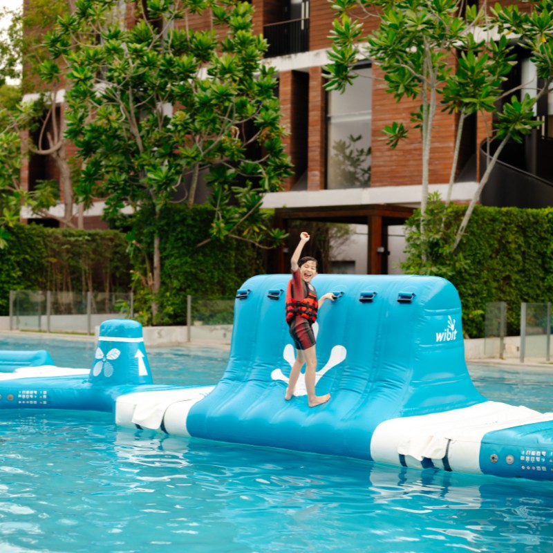 A child in a black and red swimsuit with a life vest joyfully climbs a blue inflatable in a pool. Lush greenery and a modern building backdrop.