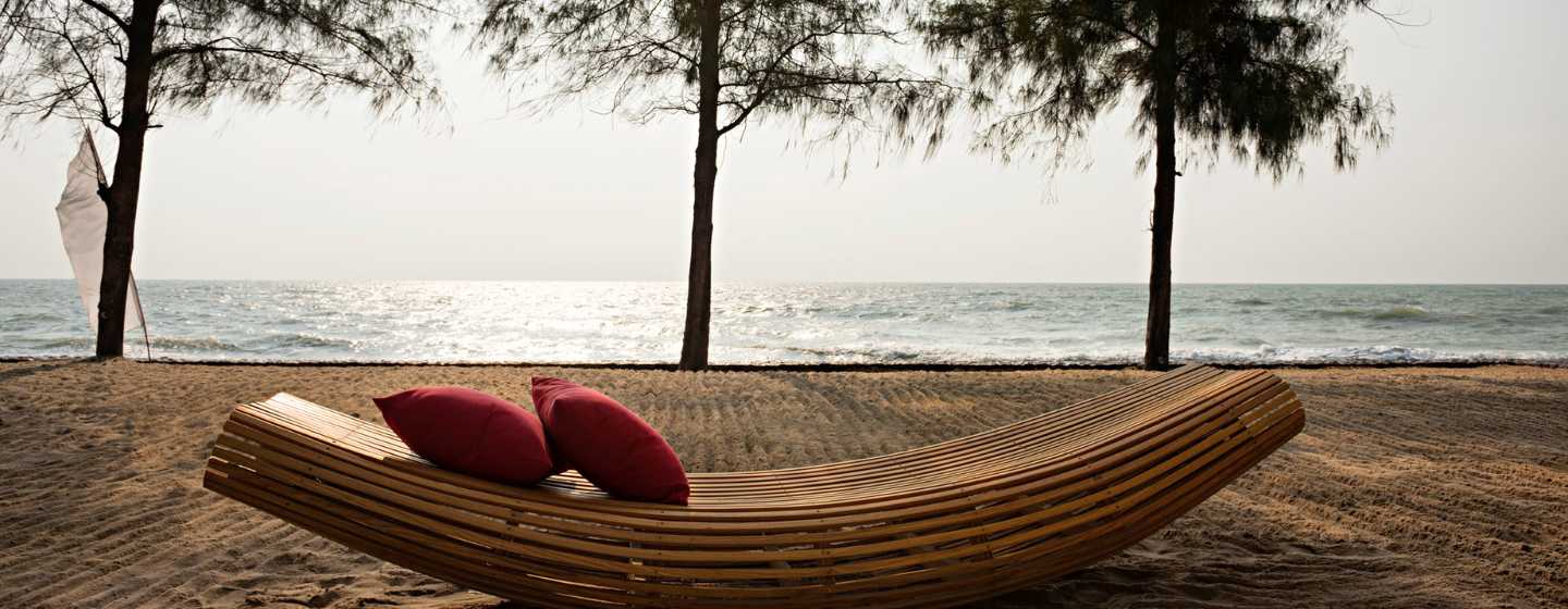Curved wooden bench with red cushions on a sandy beach, facing a calm sea. Tall trees frame the serene, overcast setting.
