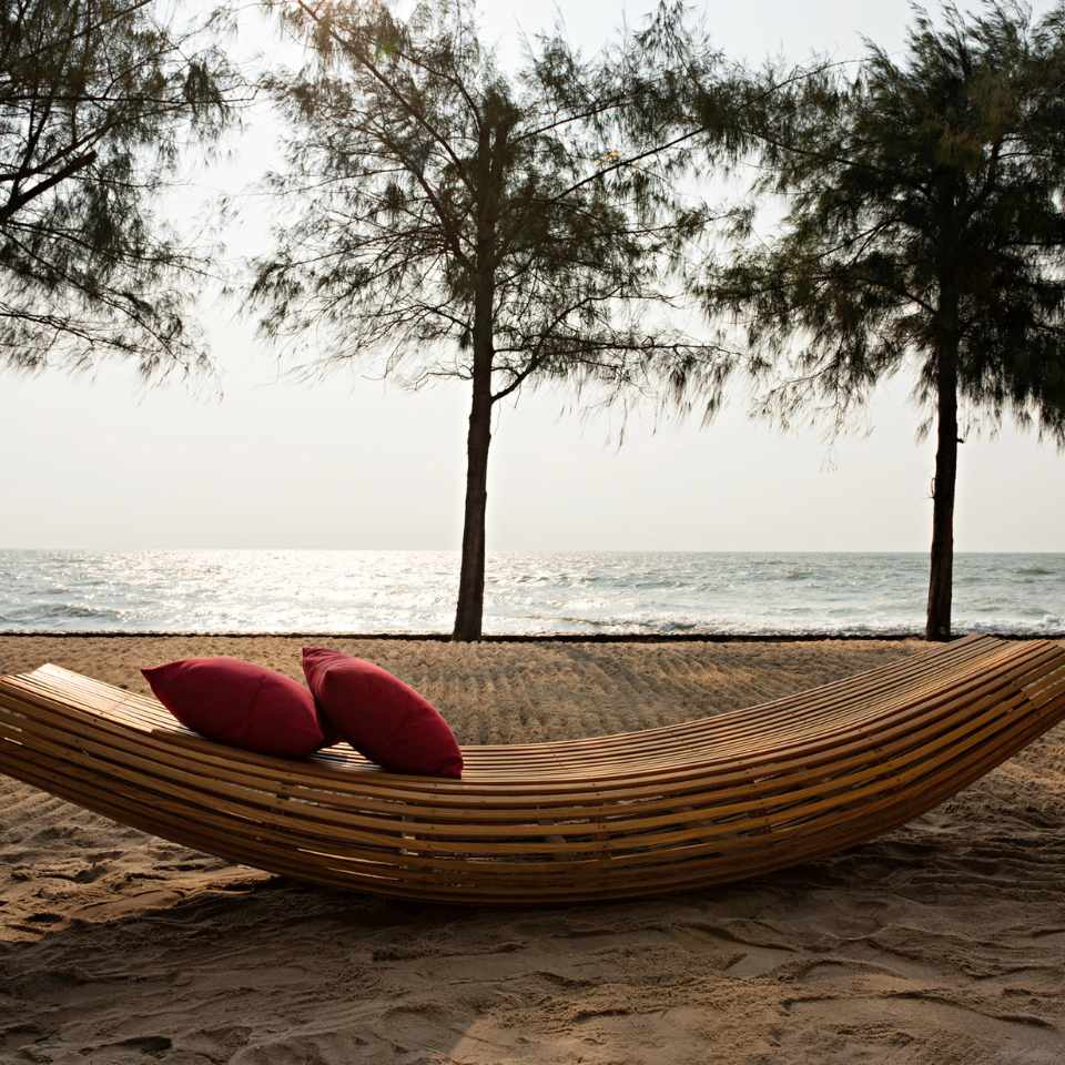 Curved wooden bench with red cushions on a sandy beach, facing a calm sea. Tall trees frame the serene, overcast setting.