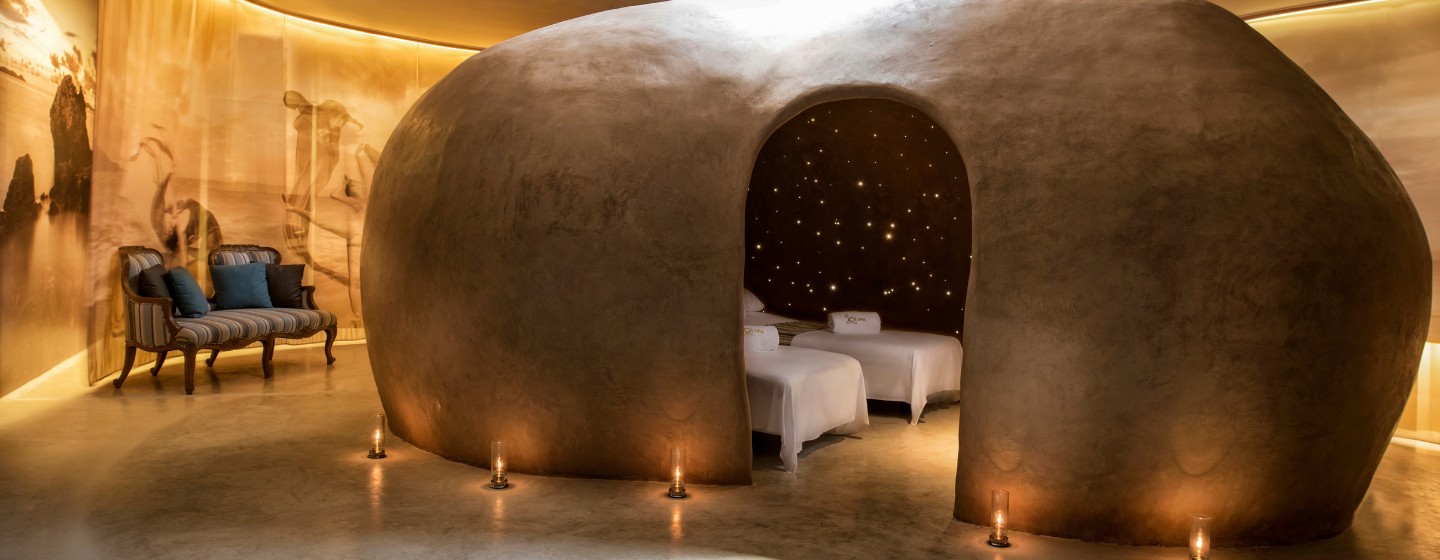 A cozy spa room features a large, oval-shaped clay structure with an archway entrance. Inside, twinkling lights resemble stars, creating a serene ambiance. Two massage beds are visible, surrounded by soft candlelight. The room has warm, earthy tones, enhancing the peaceful, relaxing atmosphere.
