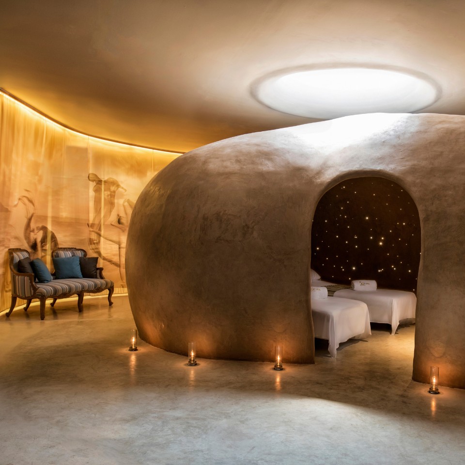 A cozy spa room features a large, oval-shaped clay structure with an archway entrance. Inside, twinkling lights resemble stars, creating a serene ambiance. Two massage beds are visible, surrounded by soft candlelight. The room has warm, earthy tones, enhancing the peaceful, relaxing atmosphere.