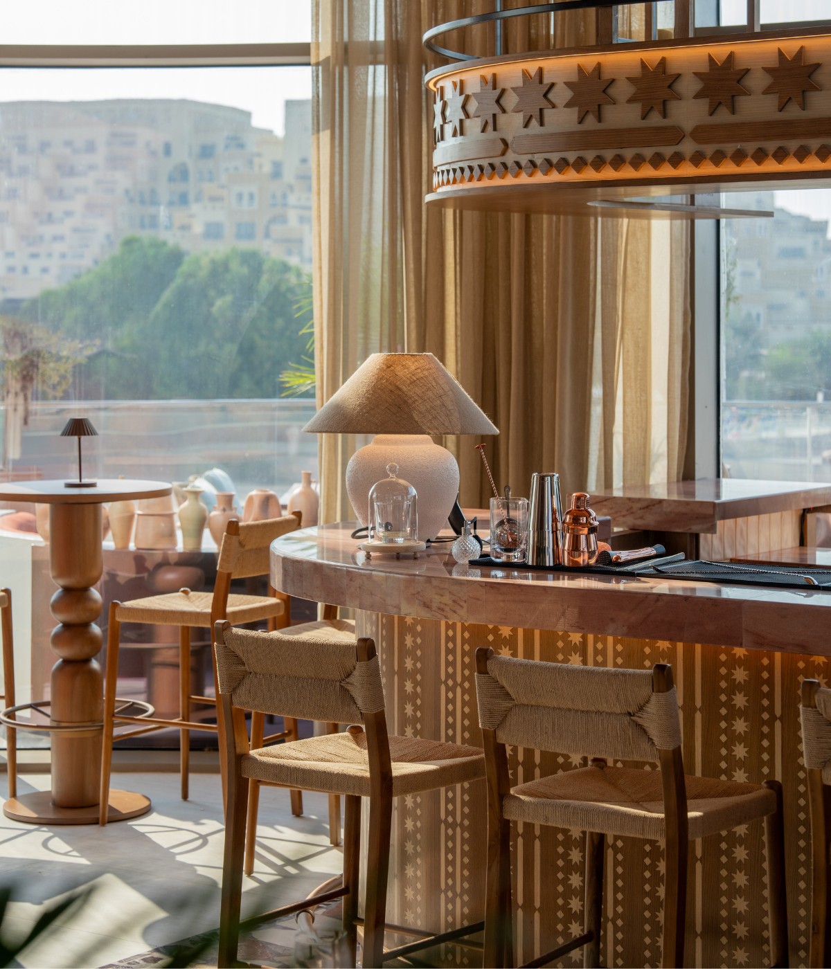 Cozy cafe interior with wooden decor, sunlight streaming through sheer curtains. A round table features a stylish lamp and bar items, creating a warm ambiance.