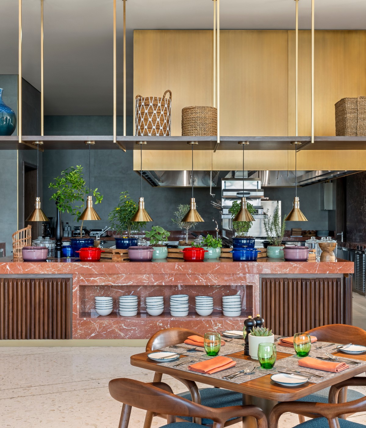 Modern kitchen with a pink marble island, colorful cookware, potted herbs, and golden pendant lights. A table with green glasses and orange napkins.
