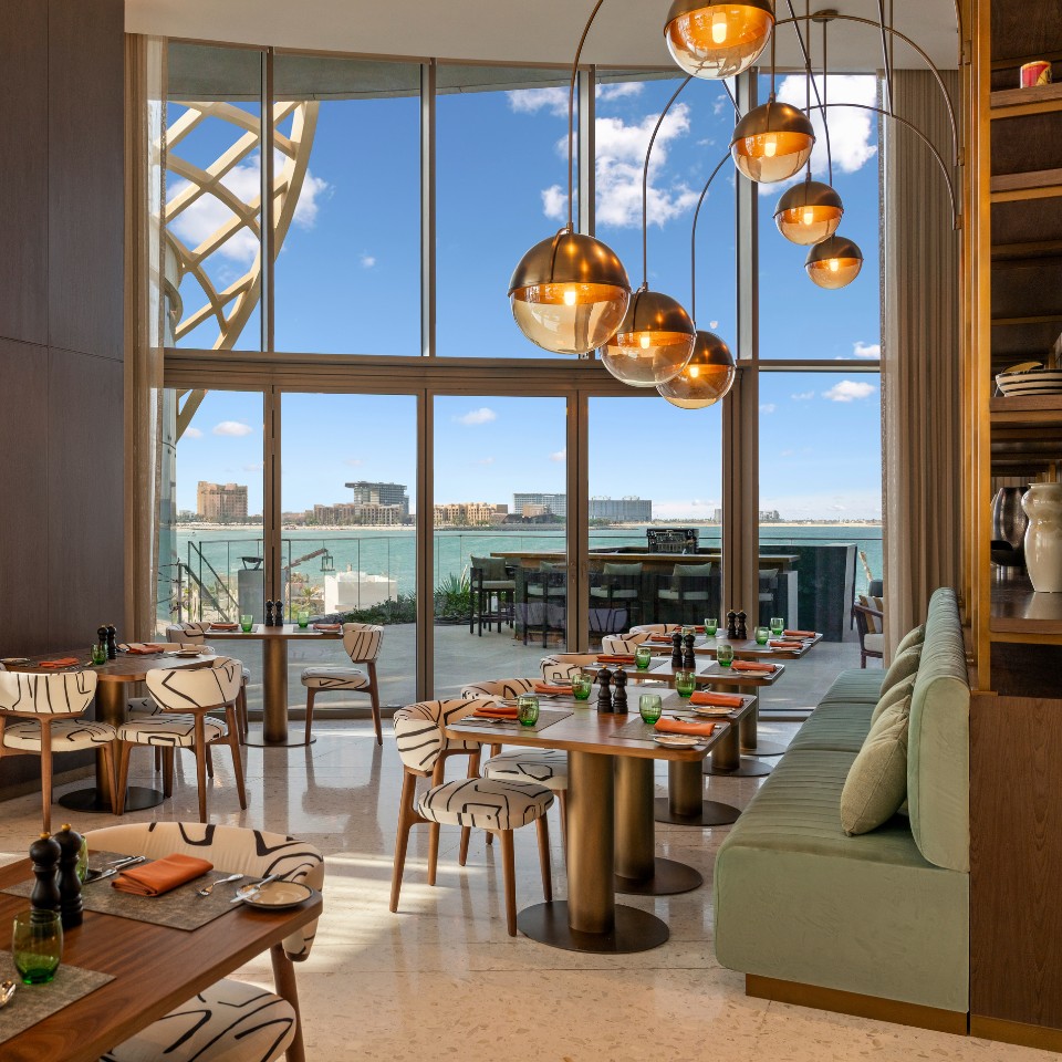 Modern restaurant with large windows offering ocean views. Tables feature elegant settings, green accents, and warm lighting, creating an inviting ambiance.