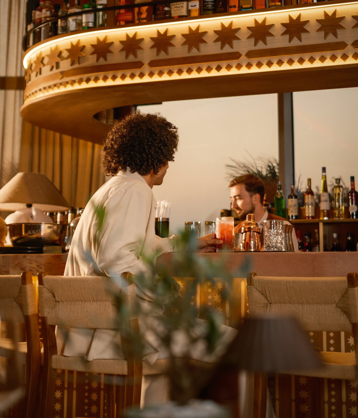 A relaxed bar scene at sunset, with a man in a light suit enjoying cocktails. The bartender converses behind a stylish counter, creating a warm, inviting atmosphere.