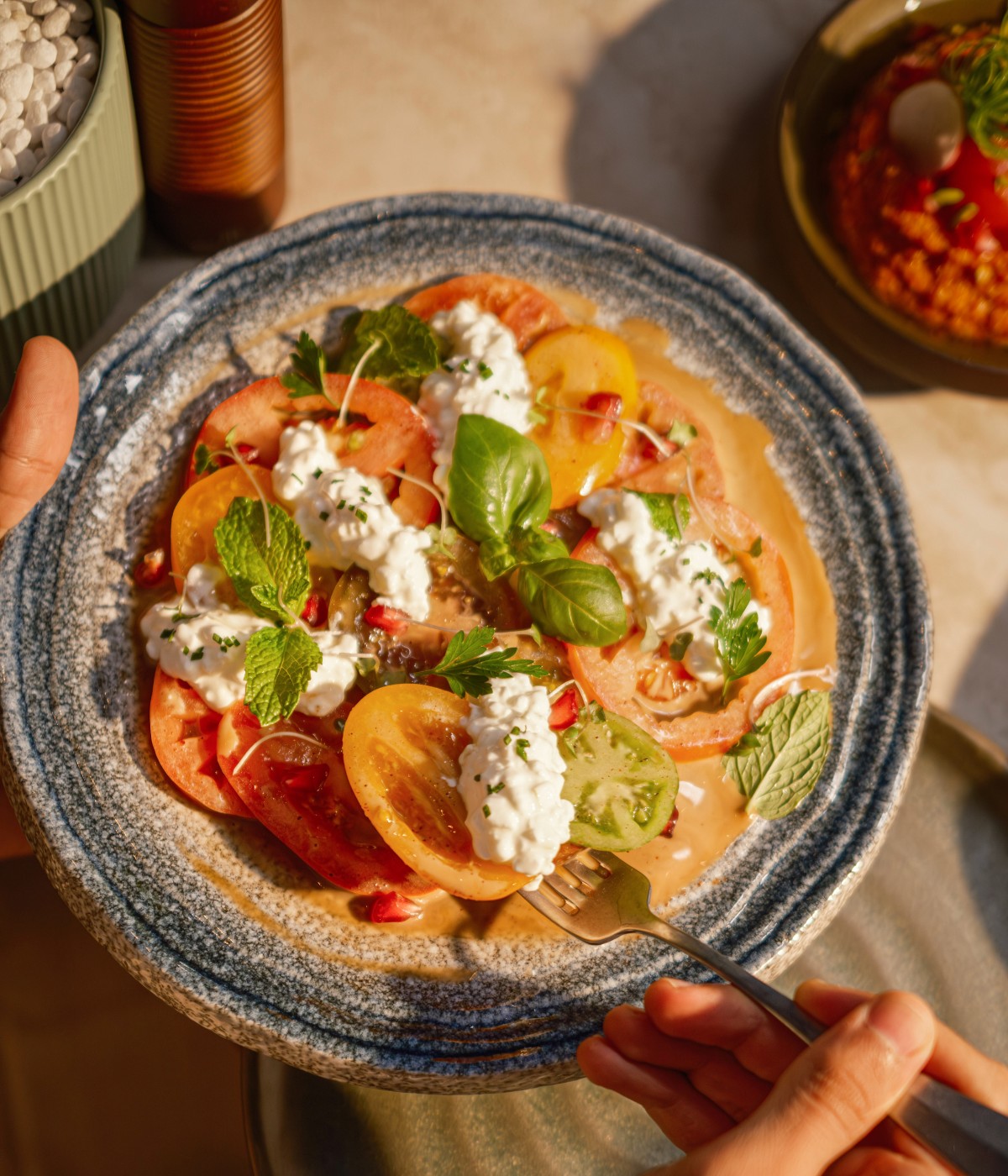 A colorful plate of heirloom tomato salad topped with creamy cheese, fresh basil, and mint, on a blue ceramic dish. A fork is poised to take a bite.