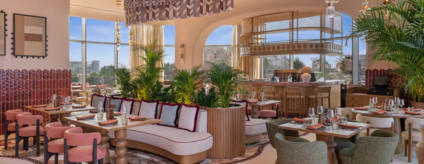 Modern restaurant interior with pastel and earthy tones, featuring plush seating, wooden tables set with glasses, lush plants, and large windows with a city view.