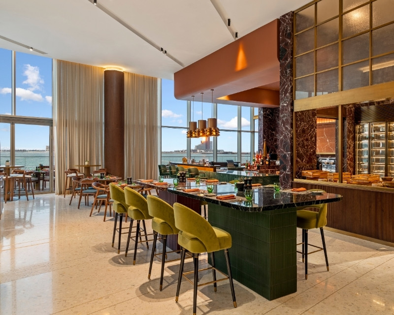http://Upscale%20restaurant%20interior%20with%20a%20modern%20design.%20Features%20green%20bar%20stools,%20a%20marble%20countertop,%20and%20large%20windows%20revealing%20a%20coastal%20view.%20Warm%20lighting%20creates%20an%20inviting%20ambiance.