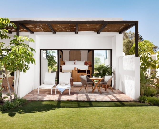 http://Modern%20patio%20with%20white%20loungers,%20small%20table,%20and%20chairs%20on%20a%20brick%20terrace.%20Open%20sliding%20doors%20reveal%20a%20cozy%20bedroom.%20Lush%20greenery%20surrounds%20the%20space.