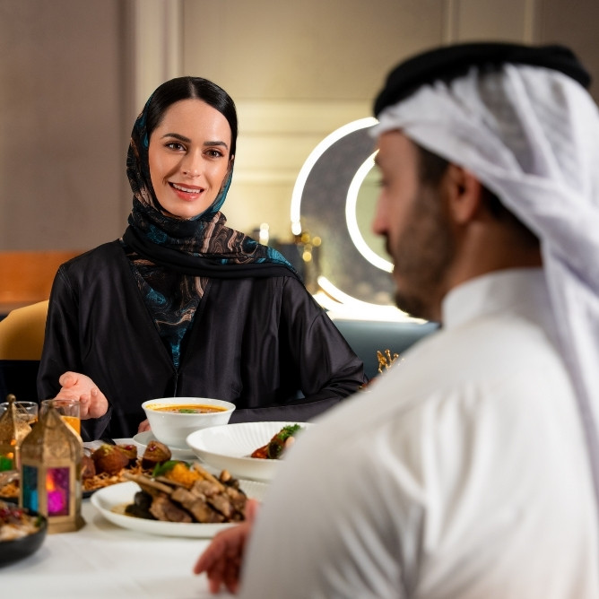 Two individuals are seated at a dining table adorned with various traditional dishes and a bowl of soup, engaged in conversation.